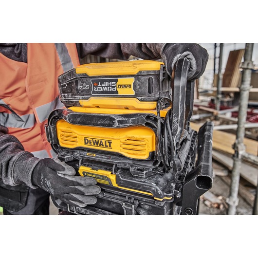 Close-up of a person wearing gloves and a safety vest, handling stacked DEWALT PowerShift 5.5Ah battery and ToughSystem storage units on a construction site.