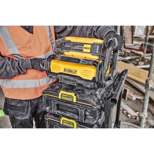 A person wearing a reflective safety vest and gloves is handling a DEWALT ToughSystem stack, which includes DEWALT PowerShift battery modules and storage boxes. The scene appears to be on a construction site.