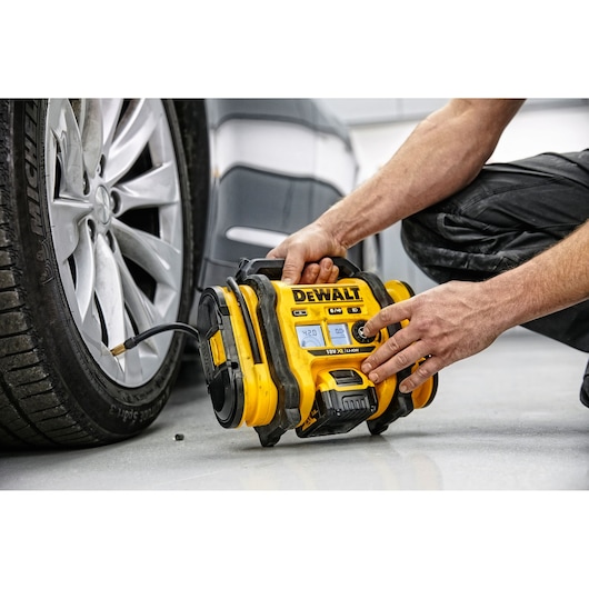 A person uses a DEWALT DCC018N portable air compressor to inflate a car tire in a garage setting. The device is yellow and black with a digital display, connected to the tire via a hose.