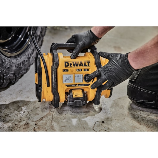 A person wearing black gloves operates a yellow DEWALT cordless portable air compressor (model DCC018N) with digital display, shown inflating a vehicle tire on a concrete surface.