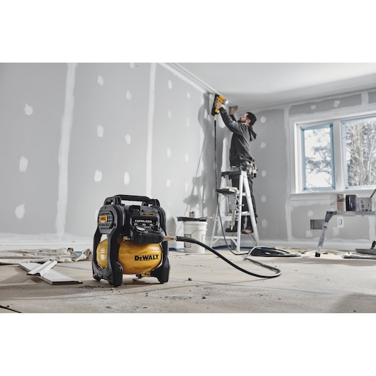 A DEWALT cordless air compressor is placed on the floor in the foreground, with a person standing on a ladder using a power tool to work on the ceiling trim in a room under construction.