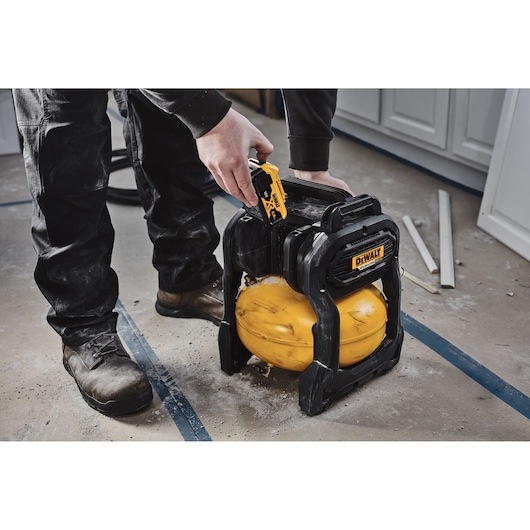 A person is using a DEWALT portable air compressor, model DCC1018N, on a construction site. The compressor is yellow and black and is placed on a dusty floor with some wooden boards nearby.