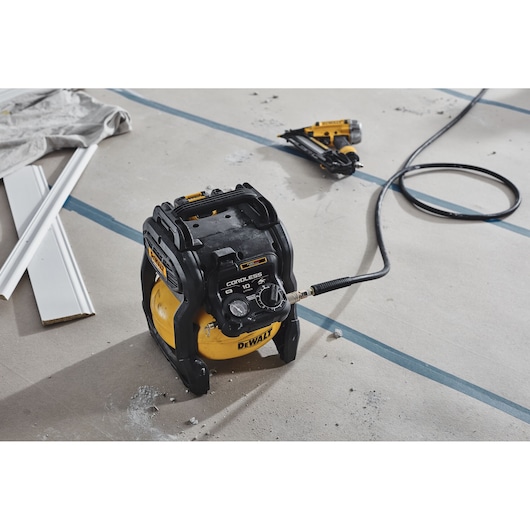 A DEWALT cordless air compressor with SKU DCC1018N is placed on a construction site floor, connected to a pneumatic tool. Some construction materials and debris are visible nearby.