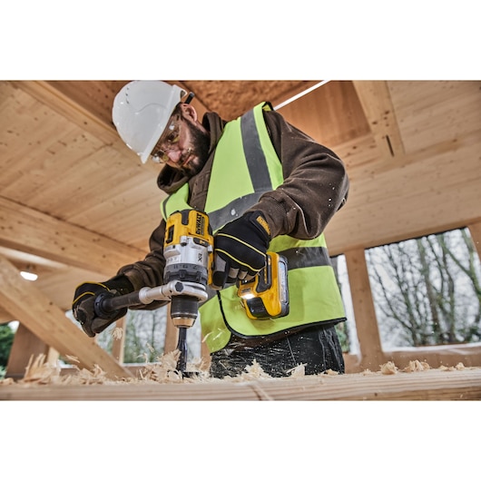 A person wearing a safety helmet, high-visibility vest, and gloves is using a DEWALT cordless drill to work on a wooden surface in a construction environment. Wood shavings are visible as the drill operates.