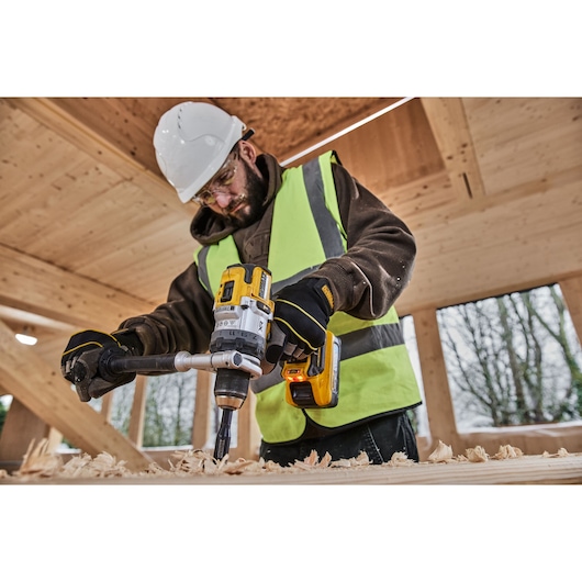 A person wearing a safety helmet and high-visibility vest uses a DEWALT cordless drill (model DCD1007H2T) to drill into a piece of wood inside a wooden construction site.