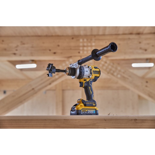 A DEWALT cordless drill with a side handle and a hole saw attachment, placed on a wooden beam in a workshop setting.