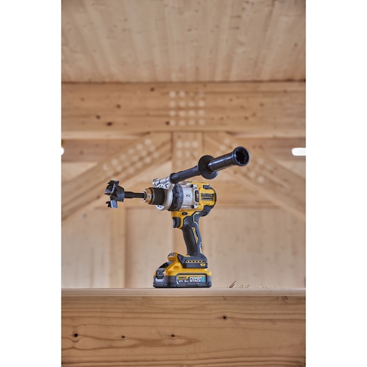 A DEWALT cordless drill (SKU: DCD1007H2T) with an attached auger bit and side handle, placed on a wooden beam in a woodworking workshop.