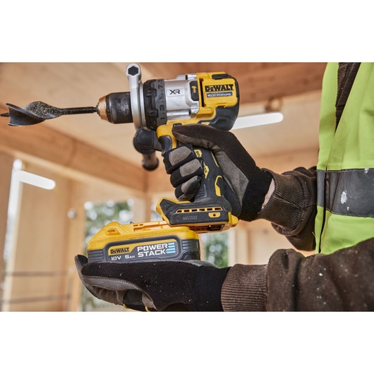 A person wearing work gloves and a high-visibility vest is holding a DEWALT cordless drill with a large auger drill bit attached. The drill features a DEWALT POWERSTACK 18V 5Ah battery. The background shows a wooden construction site.