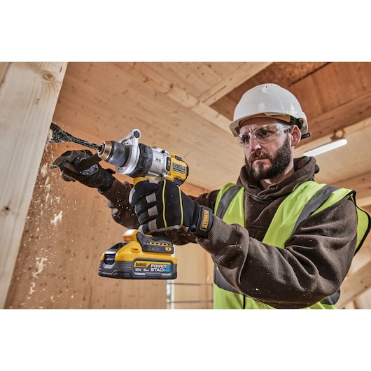 A person wearing a high-visibility vest, gloves, and a hard hat is using a DEWALT cordless drill (model DCD1007NT) to drill into a wooden beam, with wood shavings flying as the drill makes contact.