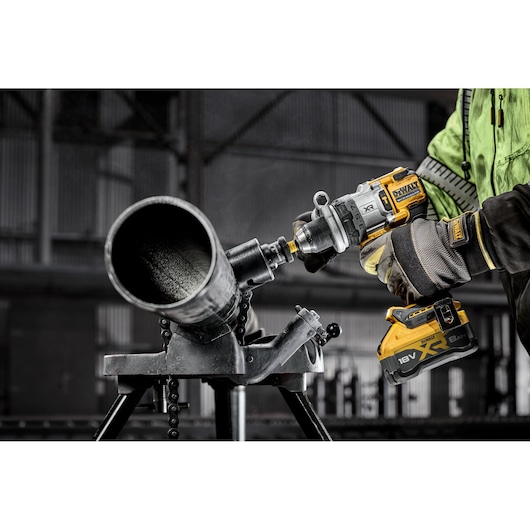 A person wearing protective gloves and a green jacket is using a DEWALT 18V XR cordless drill to drill into a large metal pipe, which is secured on a workbench in an industrial setting.