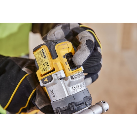 Close-up of a person wearing protective gloves, adjusting a yellow and black DEWALT cordless power tool, likely a drill or impact driver, in a workshop setting.