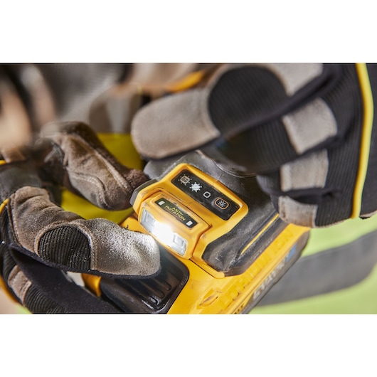 Close-up of a person wearing protective gloves about to operate the built-in light on a yellow DEWALT power tool, showing the illuminated control panel with buttons for light settings.