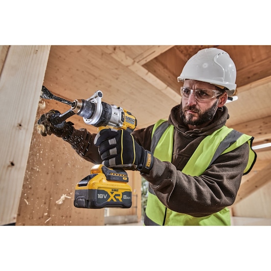 A person wearing a safety helmet, high-visibility vest, and gloves using a DEWALT DCD1007WW1T 18V XR cordless drill with a wood boring bit, drilling into a wooden beam on a construction site. The person's face is obscured.