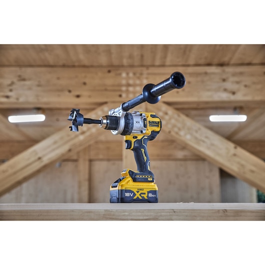 A DEWALT 18V XR cordless drill with an attached auger bit and side handle, positioned on a wooden surface in a workshop setting.