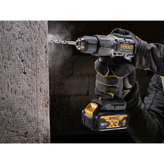 A person wearing gloves is using a DEWALT 18V XR brushless cordless drill to drill into a concrete wall. Dust is visible around the drilling point.