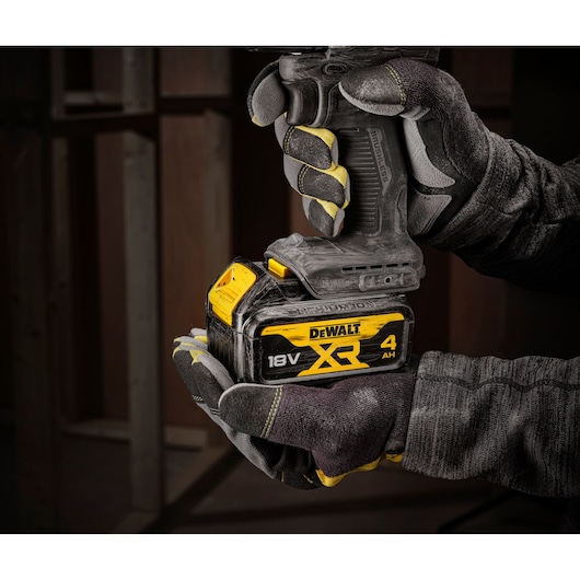 A person wearing work gloves holds a DEWALT DCD100M1T cordless power tool with an 18V XR 4Ah battery, shown in an indoor construction setting.