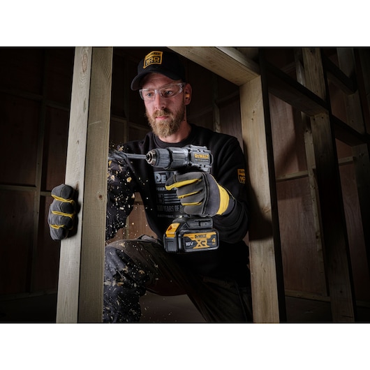 A person wearing gloves and branded workwear is using a DEWALT DCD100M2T cordless drill to drill into a wooden beam, causing wood chips to fly. The scene takes place inside a wooden frame construction.