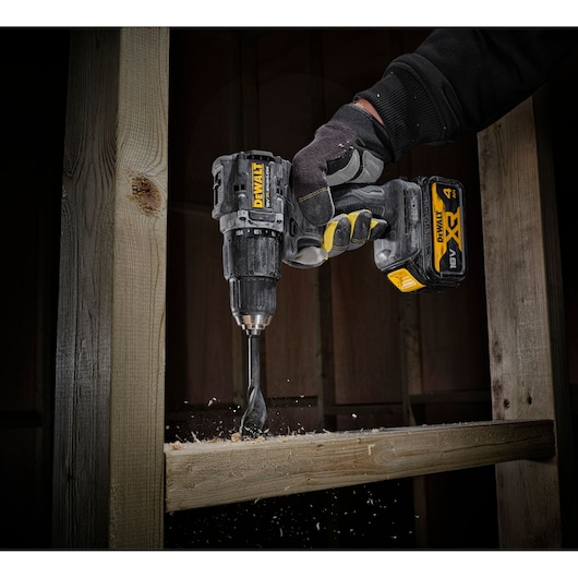 A DEWALT cordless drill is being used by a gloved hand to drill into a wooden beam, with wood shavings flying.