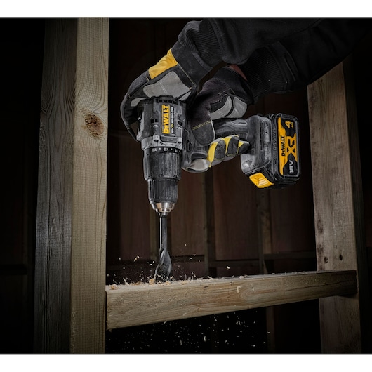 A DEWALT DCD100M2T cordless drill is being used by a gloved hand to drill into a piece of wood, with wood shavings and dust flying around.