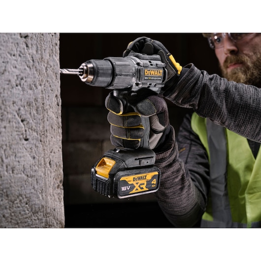 A person wearing gloves and a safety vest is using a DEWALT 18V XR cordless drill to drill into a concrete wall. The drill features a 4 Ah battery pack and is designed for heavy-duty use.