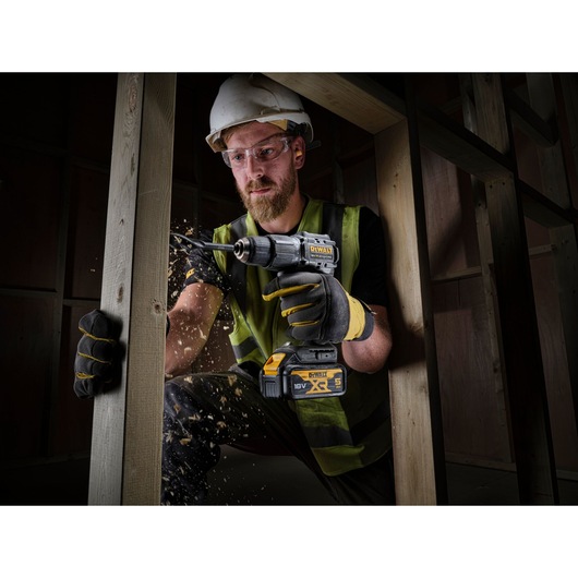 A person wearing a hard hat and safety vest is using a DEWALT 18V cordless drill to drill into a wooden beam, with wood shavings flying. The drill has a visible DEWALT logo and 5 Ah battery. The setting appears to be a construction site.