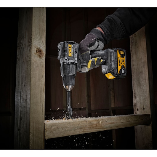 A DEWALT cordless power drill with a spade bit attached is being used by a gloved hand to drill into a wooden beam, with wood shavings flying from the drilled area.