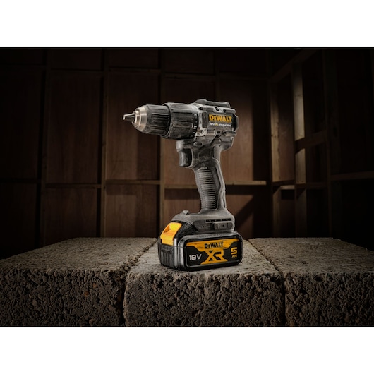A DEWALT 18V XR cordless drill with a 5.0Ah battery, standing upright on concrete blocks in a construction setting with a wooden framework in the background.