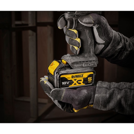A close-up view of a gloved hand holding a DEWALT 18V XR cordless tool with a 5 Ah battery, set against a workshop background.