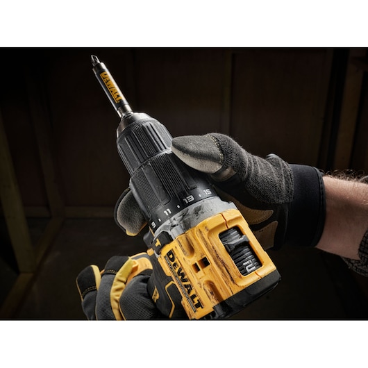 A close-up image of a gloved hand holding a DEWALT cordless power drill with a drill bit attached. The drill has a yellow and black design, and settings are visible on the front.
