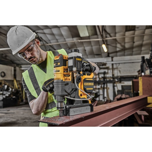 A worker wearing a safety helmet and high-visibility vest is using a DEWALT brushless magnetic drill on a steel beam inside an industrial workshop.