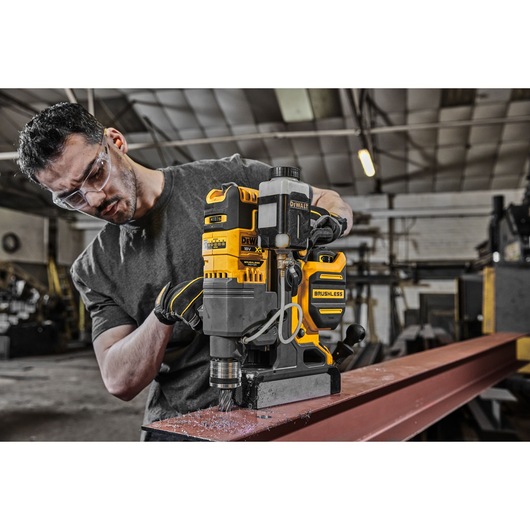 A person using a DEWALT DCD1623N brushless magnetic drill on a steel beam in an industrial workshop. The person's face is blurred. The drill is yellow and black, and metal shavings are visible around the drilling area.