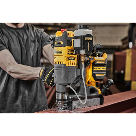 A DEWALT DCD1623N brushless magnetic drill being used to drill into a steel beam, operated by a person wearing work gloves.