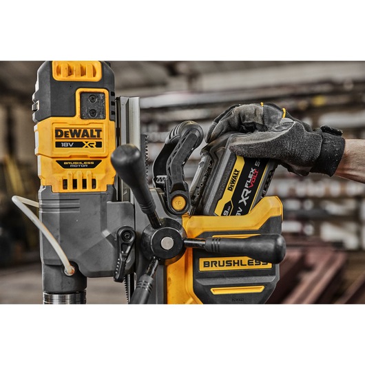 Close-up of a DEWALT 18V XR brushless motor power tool, with a gloved hand inserting a battery. The scene is indoors, in a workshop environment.