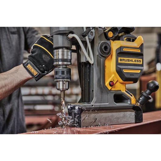 A person using a DEWALT brushless magnetic drill press to drill into a metal surface. Metal shavings are visible around the drill bit, and the person is wearing a black DEWALT glove.