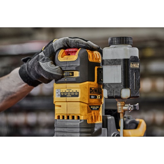 A close-up of a DEWALT 18V XR brushless motor tool with a gloved hand adjusting the settings on the top. The tool features a yellow and black design and has a liquid container attached on the side.