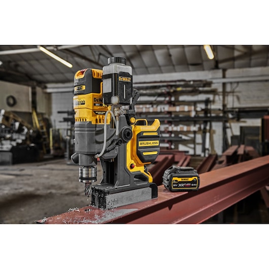 A DEWALT cordless brushless magnetic drill press with battery, shown mounted on a steel beam in an industrial workshop.