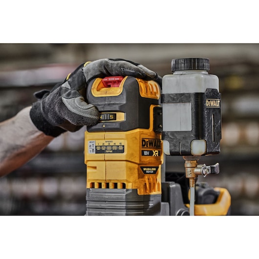 A close-up of a person wearing work gloves operating a DEWALT 18V XR brushless motor tool, model DCD1623, with a lubricant container attached on the right side.