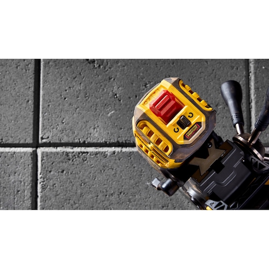 Close-up view of the top of a DEWALT power tool, showing a yellow and black housing with a prominent red button and control features, set against a textured concrete tile background.