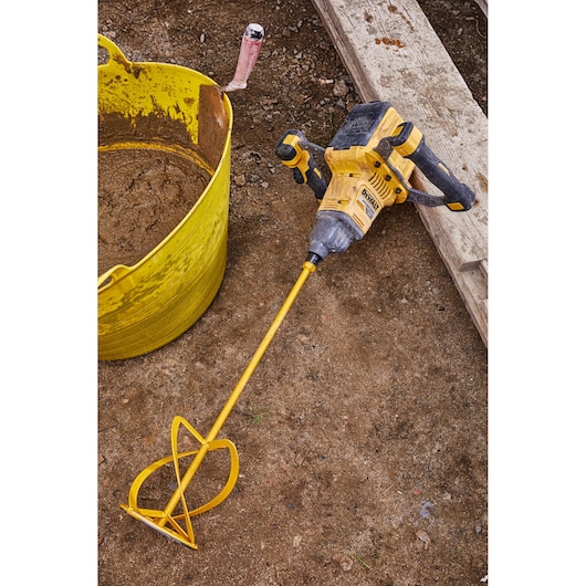 A DEWALT cordless mixer tool with a long mixing paddle attached, placed on a construction site next to a yellow tub containing mixed material.