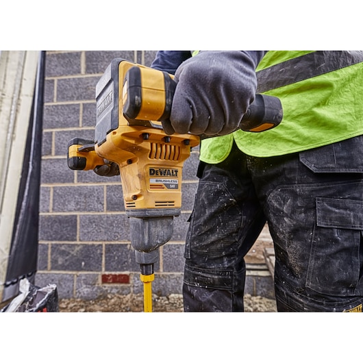 A person wearing gloves is using a DEWALT DCD240X2 brushless power drill to drill into the ground at a construction site.