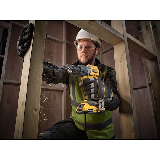 A person wearing gloves, a high-vis vest, and a hard hat is using a yellow DEWALT cordless drill (DCD799D2T) to drill into a wooden beam inside a construction site. Wood shavings are visible as the drill penetrates the wood.