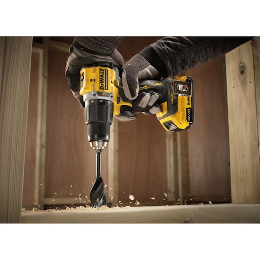 A person wearing gloves is using a DEWALT cordless drill to bore a hole in a wooden surface, with wood shavings visible.