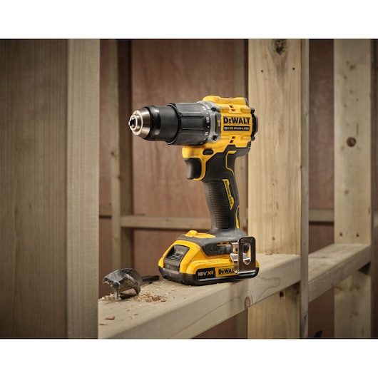 A DEWALT cordless power drill model DCD799D2T is placed on a wooden beam at a construction site. Wood shavings and a drill bit are also visible on the beam.