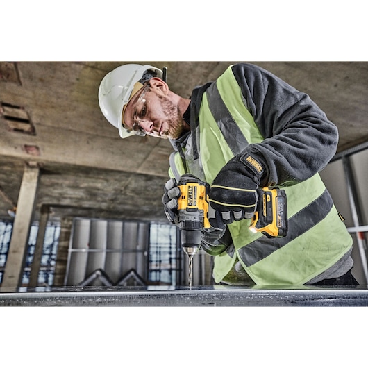 A person wearing a high-visibility vest, gloves, and a safety helmet is using a DEWALT DCD800E1T cordless drill to drill into metal at a construction site.
