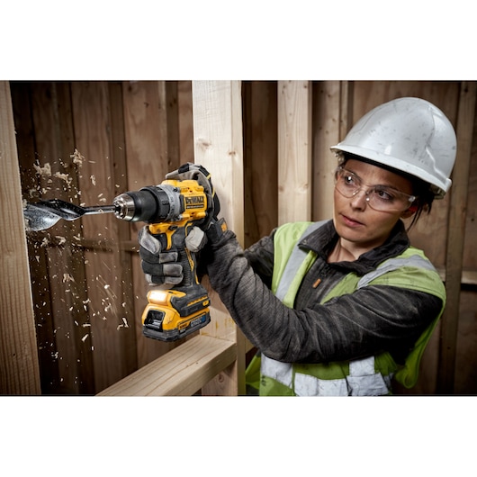 A person wearing a hard hat and safety vest is using a DEWALT cordless drill to bore a large hole through a wooden beam. Wood shavings are flying as the tool drills through the material.