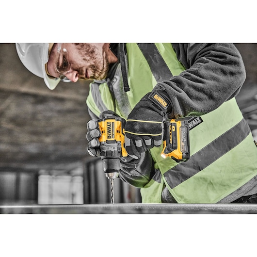 A construction worker wearing safety gear is using a DEWALT cordless drill (model DCD800E2T) to drill into a metal surface in an industrial setting.