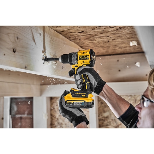 A person wearing safety glasses and gloves is using a DEWALT cordless drill to bore a hole in a wooden beam, with wood shavings flying during the process.