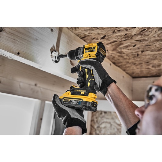 A person wearing gloves uses a DEWALT cordless drill to drill a hole into a wooden beam in a construction setting.