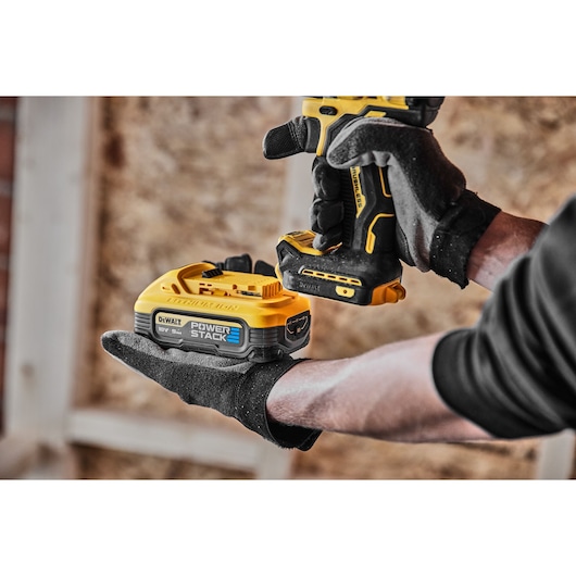 A person wearing black gloves is holding a DEWALT 18V POWERSTACK battery and a DEWALT cordless power tool inside a construction area with wooden walls.