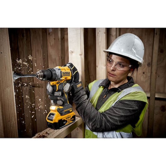 A construction worker wearing a safety helmet and vest is using a DEWALT DCD800P2T cordless drill to bore a hole into a wooden beam. Wood shavings are flying from the drilling site.